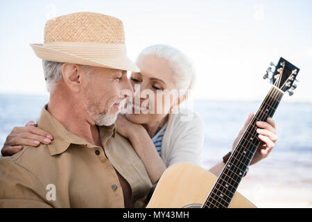 Felice senior abbracciando la donna uomo barbuto in hat a suonare la chitarra all'aperto Foto Stock