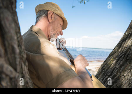 Senior uomo in hat poggiando su albero e a suonare la chitarra al Riverside Foto Stock