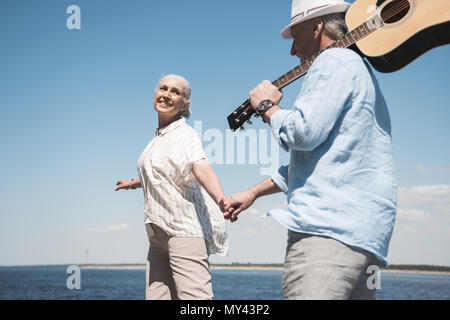 Felice coppia senior con la chitarra tenendo le mani e piedi al quay Foto Stock