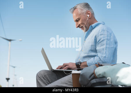 Uomo anziano lavora su laptop e ascoltando musica in earpods all'aperto Foto Stock