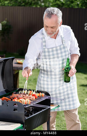 Capelli grigi uomo beve birra dalla bottiglia durante la preparazione di barbecue sul grill. Foto Stock