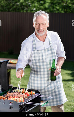 Sorridente dai capelli grigi uomo beve birra dalla bottiglia durante la preparazione di barbecue sul grill Foto Stock