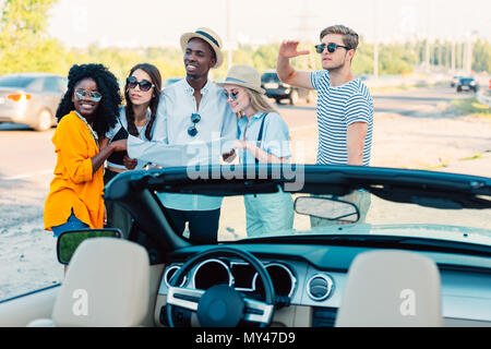 Gruppo multietnico di amici a utilizzare la mappa per determinare la direzione mentre sta in piedi vicino a vettura insieme Foto Stock