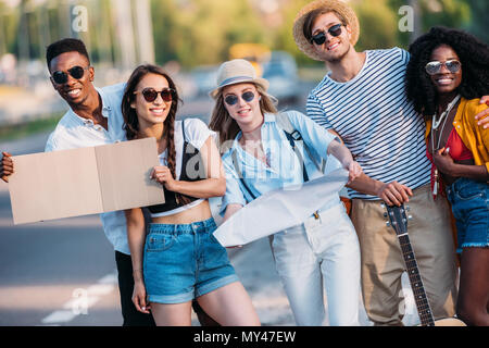 Gruppo multiculturale dei giovani hitchhikers con mappa e cartone vuoto permanente al marciapiede della strada Foto Stock