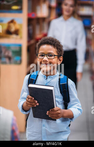 Giovani African American boy holding libro in biblioteca con docente Foto Stock