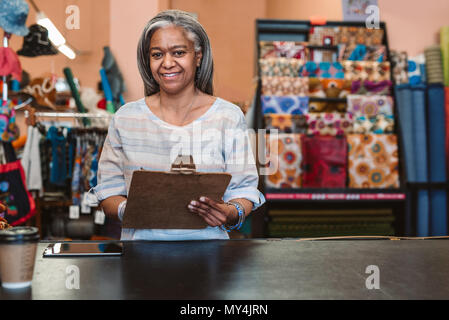 Ritratto di un sorridente tessuto maturo proprietario del negozio in piedi dietro un contatore Circondato da colorati tessuti e prodotti tessili e tenendo un clipboard Foto Stock