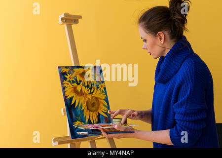 Ragazza artista dipinge i girasoli Vernici a olio su tela. Indossa un maglione blu. La donna è in possesso di pennello e la tavolozza dei colori con vernici. Lei mescola di colori Foto Stock