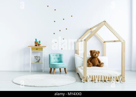 Orsacchiotto di peluche sul letto di legno accanto alla sedia blu in bianco di capretto interiore camera da letto. Foto reale Foto Stock