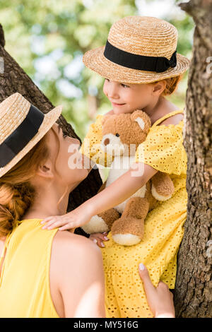 Giovane madre holding incantevole piccola figlia con orsacchiotto seduto su albero a park Foto Stock