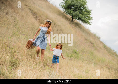 Felice bambina e la giovane donna con la valigia tenendo le mani e piedi sulla collina erbosa Foto Stock