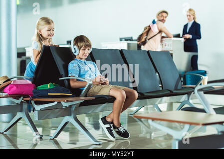 Ragazzo in cuffie utilizzando smartphone con la sorella vicino dal padre e al banco per il check in all'aeroporto Foto Stock