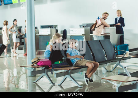 Ragazzo in cuffie utilizzando smartphone con la sorella vicino dal padre e al banco per il check in all'aeroporto Foto Stock