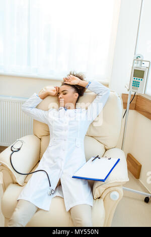 Giovane africano-medico americano giacente in poltrona esaurito con uno stetoscopio e appunti Foto Stock