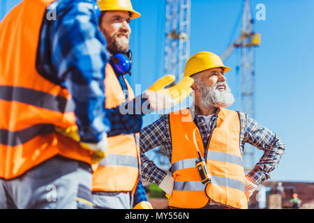 Tre costruttori in giubbotti riflettenti e hardhats discutendo di lavorare al sito in costruzione Foto Stock