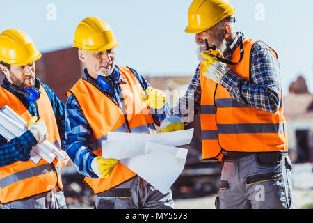 Tre lavoratori in uniforme di esaminare i progetti di un edificio e parlare su radio portatile al sito in costruzione Foto Stock