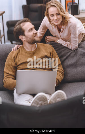 Donna sorridente guardando disabilitato marito mettere le gambe su una sedia a rotelle e utilizzo portatile a casa Foto Stock