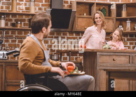 Felice madre e figlia guardando il padre seduto nella sedia a rotelle e tagliare le verdure per la cena Foto Stock