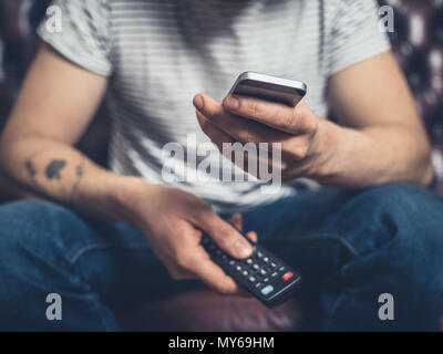 Un giovane uomo è seduto su un divano con un telecomando e uno smartphone Foto Stock