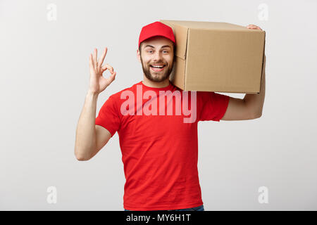 Giovane uomo di consegna nel cappuccio rosso in piedi con pacco postale box isolate su sfondo bianco che mostra bene gesto. Foto Stock