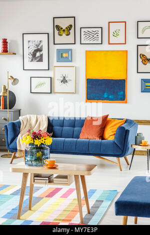 Bouquet di fresco, fiori di primavera in un vaso di vetro su un tavolo da caffè in legno nel mezzo di un luminoso soggiorno interno con divano blu e poster su Foto Stock