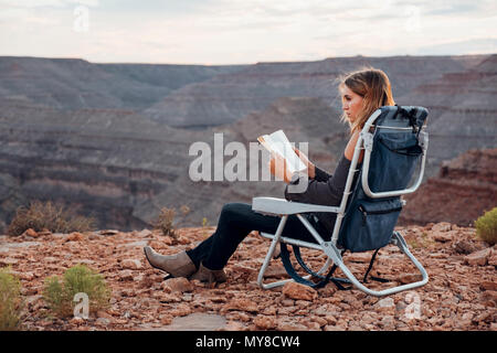 Giovane donna in impostazione remota, seduti su una sedia da campeggio, lettura libro, Mexican Hat, Utah, Stati Uniti d'America Foto Stock