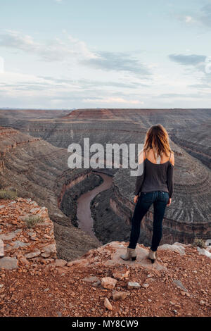 Giovane donna in impostazione remota, in piedi sul bordo di scogliera, guardando a vista, vista posteriore, Mexican Hat, Utah, Stati Uniti d'America Foto Stock