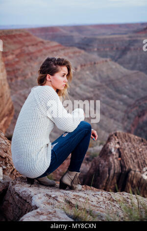 Giovane donna in impostazione remota, seduti sulle rocce, guardando a vista, Mexican Hat, Utah, Stati Uniti d'America Foto Stock