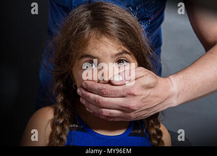 La mano ruvida di un uomo adulto chiude il palmo della bocca di una bambina con occhi spaventati contro una parete scura Foto Stock