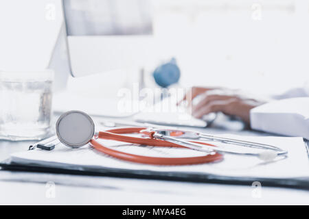 Messa a fuoco morbida dello stetoscopio rosso sul modulo di domanda o elenco di controllo del tavolo di lavoro con il medico donna handworking con il computer. Sanità e medicina con Foto Stock