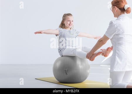 Sorridente ragazza seduta sulla grande palla lavora con fisioterapista Foto Stock