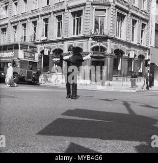 1960, storico, diurno e una uniforme della British poliziotto con hemet è visto dirigere traffico nel mezzo di una strada di Londra, Inghilterra, Regno Unito. Foto Stock