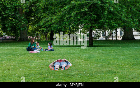 Un uomo che giace piatto su erba, completamente rilassato, donna seduta con i bambini nella zona dietro di lui, St James Park, Londra, Inghilterra, Regno Unito Foto Stock