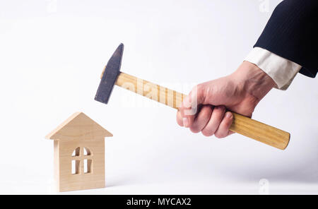 Un uomo sta colpendo con un martello una miniatura casa in legno su uno sfondo bianco. Concetto di riparazione delle abitazioni. Il lavoratore svolge lavori di riparazione su fa Foto Stock