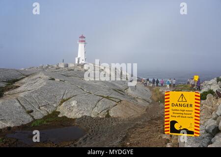 Peggy's Cove, Nova Scotia, Canada: Peggy's Point Lighthouse (1914) è avvolta nella nebbia come un segno in primo piano mette in guardia i turisti a rimanere fuori rocce umide. Foto Stock