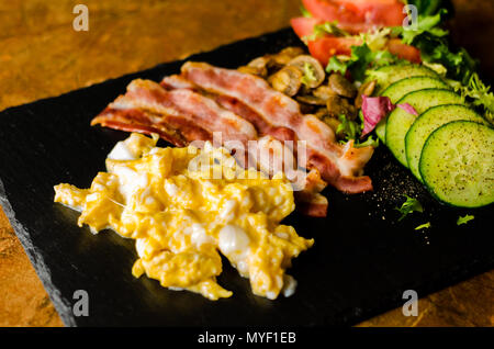 Uova strapazzate con bacon, funghi porcini servito con insalata di pomodori e cetrioli, set colazione Foto Stock