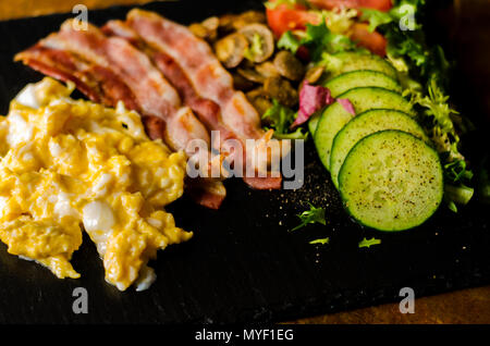 Uova strapazzate con bacon, funghi porcini servito con insalata di pomodori e cetrioli, set colazione Foto Stock