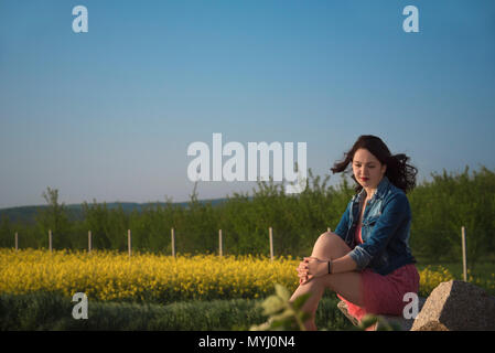 Bruna giovane donna, in un abito rosa, seduto su una roccia, nei pressi di un giallo campo di colza e un frutteto, in Moravia del Sud, Repubblica ceca, al tramonto. Foto Stock