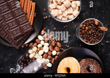 Vetro con i chicchi di caffè accanto a tavolette di cioccolato, ciambelle, lo zucchero di canna e di altro vetro con arachidi in cioccolato Foto Stock