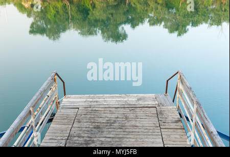 Vecchio di legno e metallo arrugginito jetty di un lago o fiume con ancora acqua e riflessioni del verde circostante alberi Foto Stock