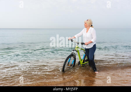 Donna bionda guadare attraverso il mare spingendo una bicicletta attraverso il surf come ella guarda sull'oceano calmo Foto Stock
