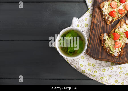 Con tè alla menta fresca in una tazza bianca su un tovagliolo e un nero lo sfondo di legno. Sana colazione toast con avocado e fragole. Spazio di copia, Foto Stock