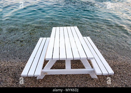 Una panca bianca da picnic è situata proprio sulla costa, con le acque cristalline turchesi che lambiscono dolcemente al bordo. Foto Stock