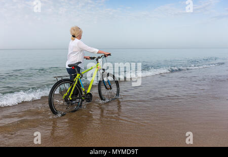 Donna che cammina lungo una spiaggia in acqua poco profonda in bordo di mare spingendo una bicicletta allontanandosi dalla fotocamera con spazio di copia Foto Stock