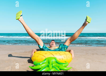 Un ridere uomo caucasico seduto su un anello di nuotare in forma di ananas sulla spiaggia, con le sue gambe sollevata, con l'oceano sullo sfondo Foto Stock
