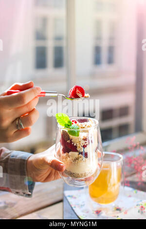 Il tè alle erbe e muesli con frutti di bosco in un bicchiere. La donna è la prima colazione. La colazione vicino alla finestra nel soggiorno. Mangiare sano. Posto per il testo. I colori della luce Foto Stock