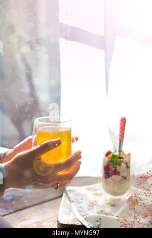 Il tè alle erbe in mani femminili. Muesli con frutti di bosco in un bicchiere. La colazione vicino alla finestra nel soggiorno. Mangiare sano. Posto per il testo. I colori chiari. Foto Stock