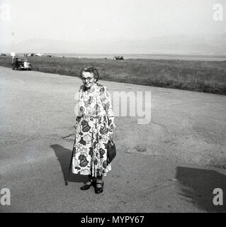 1953, Luglio, Inghilterra, foto storiche di una anziana signora inglese con bastone che indossa un abito floreale in piedi al di fuori su una strada vicino alla costa. Foto Stock