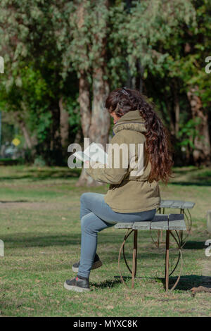 Una donna che legge un libro in una giornata di sole in un parco, seduta su una panchina Foto Stock