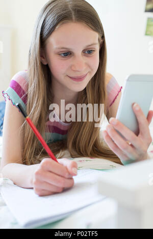 Ragazza distesa sul letto utilizzando il telefono cellulare per aiutare con i compiti Foto Stock