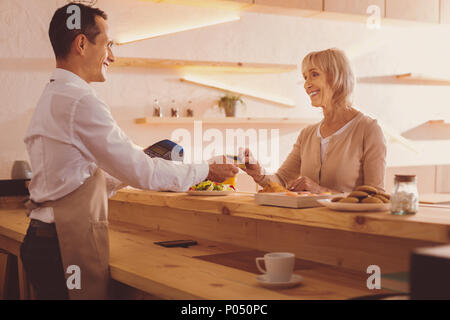 Comodo il pagamento. Piacevole donna senior dando la sua carta bancaria al barista, pagando per il cibo ordinato in cafe, mentre sorridente a lui splendidamente Foto Stock